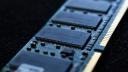 Macro close-up of a computer RAM module, showing the blue circuit board and metallic connectors in sharp detail with a shallow depth-of-field blur.
