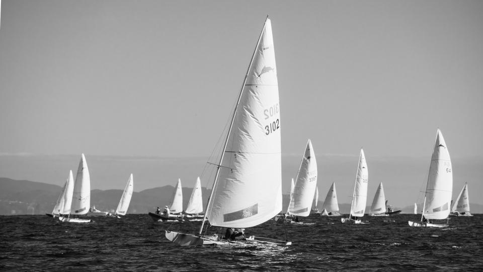 Black and white photo of a fleet of small sailboats racing on open sea, with one boat, sail number 3102, in front and hills in the background.

